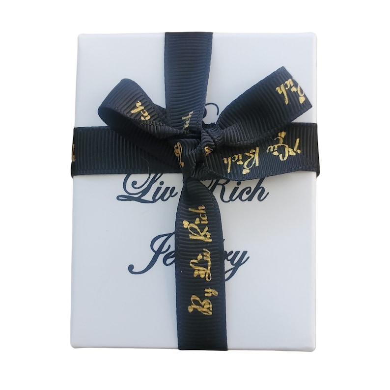 May include: A white gift box with a black ribbon tied in a bow. The ribbon has gold lettering that says "Liv Rich Jewelry" and "By Liv Rich".