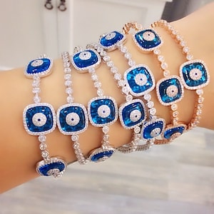 May include: A set of six silver bracelets with blue evil eye charms and clear cubic zirconia stones. The bracelets are layered on a wrist.