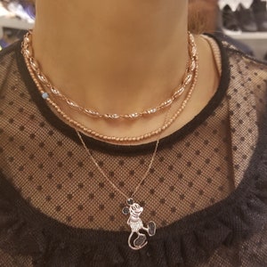 May include: A layered necklace with a rose gold finish. The top layer features a chain of small, oval-shaped beads. The middle layer is a delicate chain with a single blue bead. The bottom layer features a pendant of Mickey Mouse with black crystal accents.