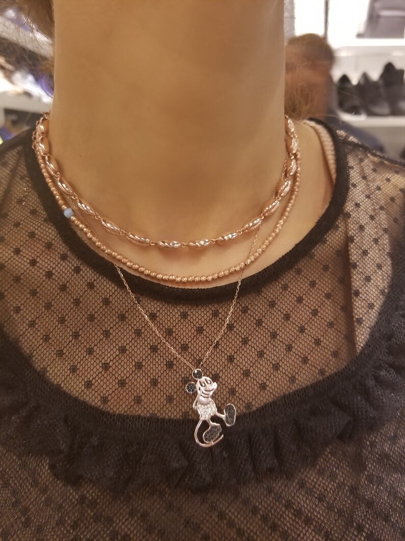 May include: A layered necklace with a rose gold finish. The top layer features a chain of small, oval-shaped beads. The middle layer is a delicate chain with a single blue bead. The bottom layer features a pendant of Mickey Mouse with black crystal accents.