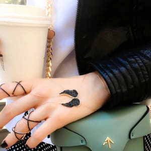 May include: A gold chain necklace with a black crystal angel wing pendant. The necklace is worn by a person with a black leather jacket and a white shirt.