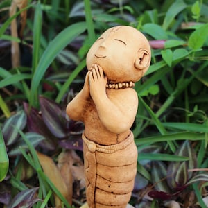 Puede incluir: Una pequeña figurita de arcilla marrón de una persona con las manos juntas en oración. La figurita lleva una larga túnica marrón y tiene una expresión serena en su rostro. La figurita está de pie sobre una base con el texto "Hare Krishna" escrito en ella.