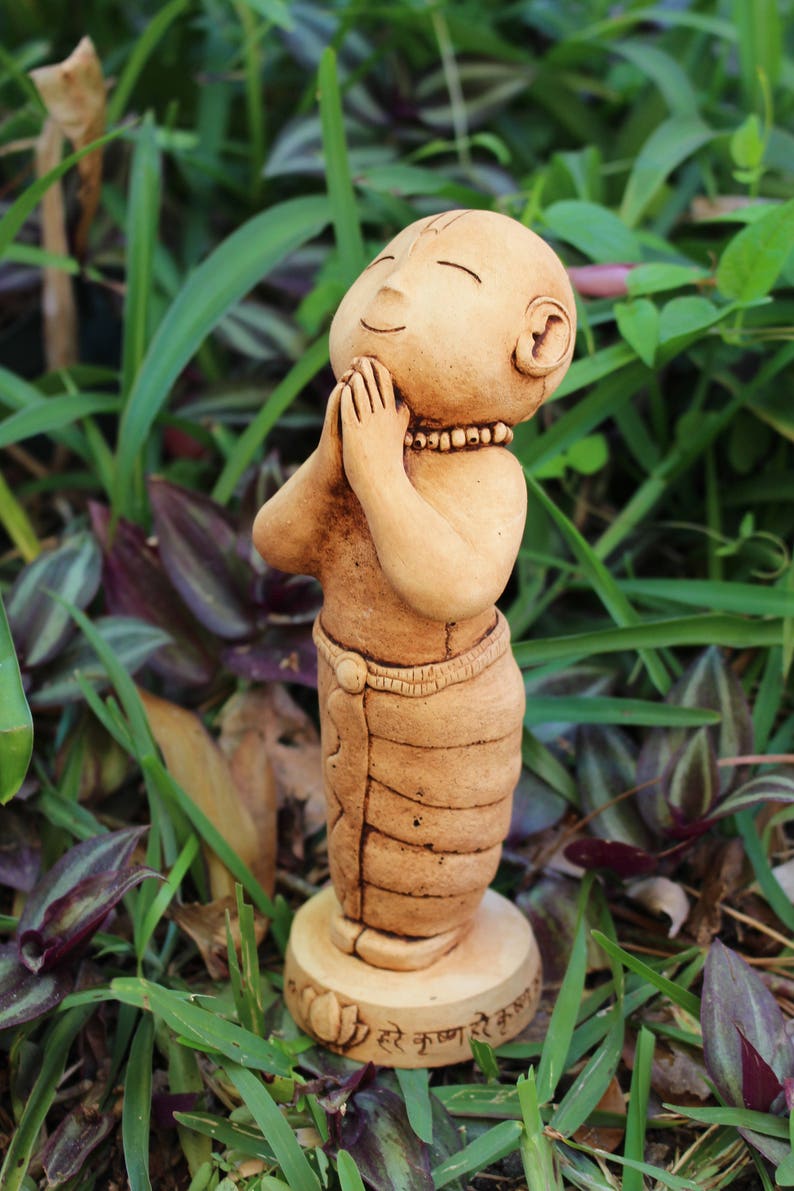 May include: A small, brown clay figurine of a person with their hands clasped in prayer. The figurine is wearing a long, brown robe and has a serene expression on its face. The figurine is standing on a base with the text "Hare Krishna" written on it.