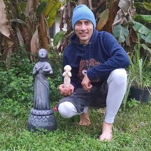 May include: A person wearing a blue beanie and a dark blue hoodie with a white and black patterned design is kneeling in a grassy area. The person is holding a small white statue in their right hand. There are two grey statues on the ground in front of the person.