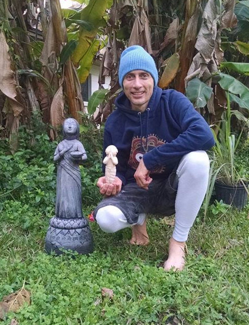 May include: A person wearing a blue beanie and a dark blue hoodie with a white and black patterned design is kneeling in a grassy area. The person is holding a small white statue in their right hand. There are two grey statues on the ground in front of the person.