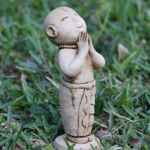 May include: A small, white, ceramic statue of a person with their hands clasped in prayer. The statue is standing on a small base and is surrounded by green grass.