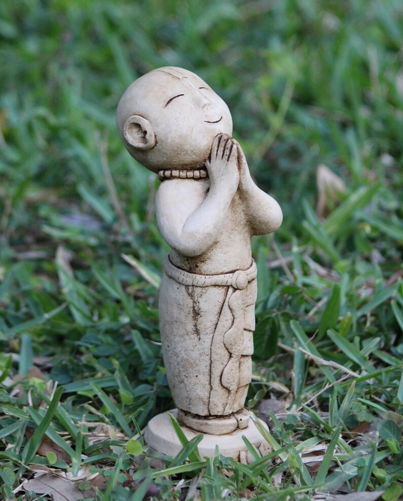 May include: A small, white, ceramic statue of a person with their hands clasped in prayer. The statue is standing on a small base and is surrounded by green grass.