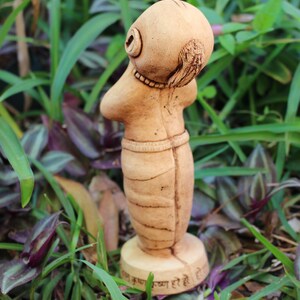 May include: A small, hand-crafted clay figurine of a person with a rounded head and a long, flowing body. The figurine is painted in a light brown colour and has a base with the text "Jai Krishna Krishna Hare Hare" inscribed on it.