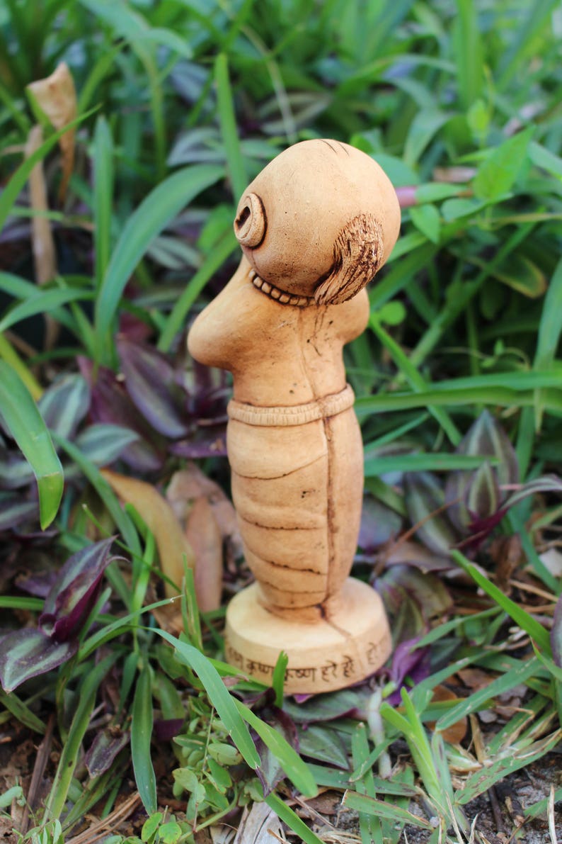 May include: A small, hand-crafted clay figurine of a person with a rounded head and a long, flowing body. The figurine is painted in a light brown colour and has a base with the text "Jai Krishna Krishna Hare Hare" inscribed on it.