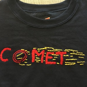 May include: A black sweatshirt with the word "COMET" embroidered in red thread. The "O" in comet is a circle with a yellow line through it. The word is followed by a trail of yellow stitches.