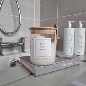 Round Glass jar with bamboo lid and spoon hung from side of jar with square white label with bath salts written in thin capital font in centre of label. Jar sat on a grey bath board over a white bath
