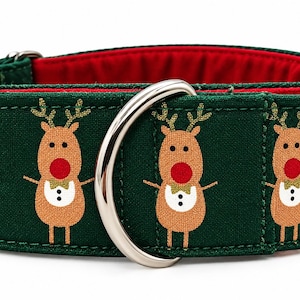 May include: A dark green pet collar featuring a repeating pattern of tan reindeer with red noses, white bibs, and gold bow ties. The collar has a bright red interior lining and a large, polished silver metal D-ring attached to the front.