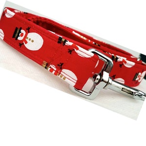 May include: A red fabric dog leash featuring a repeating pattern of white snowmen with black hats and orange noses. The leash includes a silver-toned metal swivel snap hook attached to the end of the fabric strap with reinforced stitching.