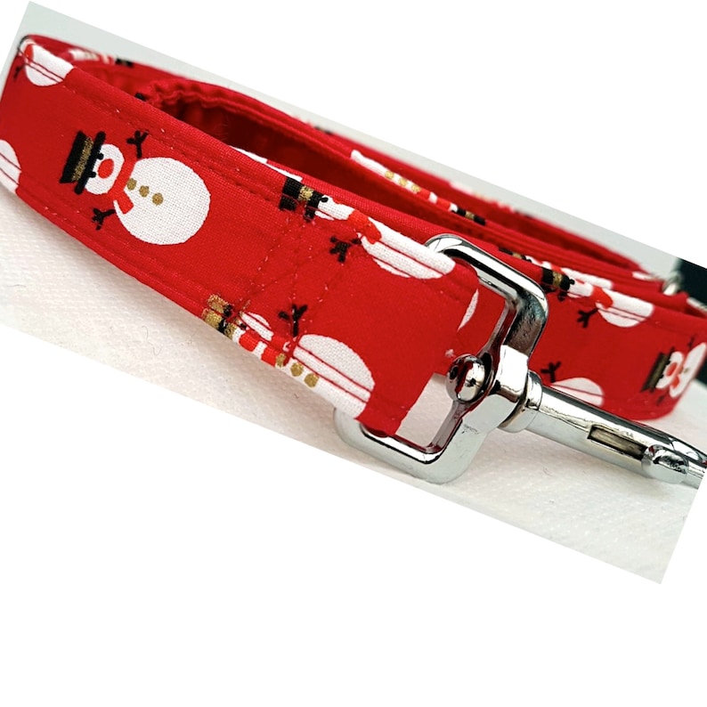 May include: A red fabric dog leash featuring a repeating pattern of white snowmen with black hats and orange noses. The leash includes a silver-toned metal swivel snap hook attached to the end of the fabric strap with reinforced stitching.