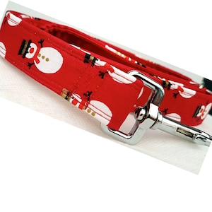 May include: A red fabric dog leash featuring a repeating pattern of white snowmen with black hats and orange noses. The leash includes a silver-toned metal swivel snap hook attached to the end of the fabric strap with reinforced stitching.