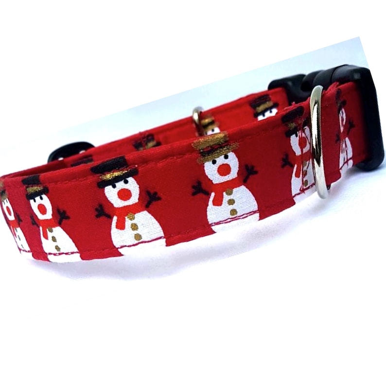 May include: A red pet collar featuring a repeating pattern of white snowmen with black top hats, orange noses, and red scarves. The collar includes a black plastic side-release buckle and a silver-toned metal D-ring for attaching a leash.