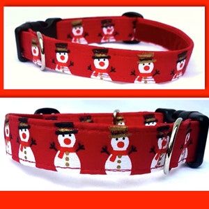 May include: A red pet collar featuring a repeating pattern of white snowmen with black top hats, orange noses, and red scarves. The collar includes a black plastic side-release buckle and a silver-toned metal D-ring for attaching a leash.