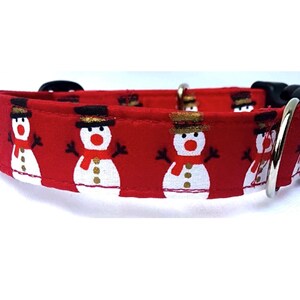 May include: A red fabric pet collar featuring a repeating pattern of white snowmen with black top hats, red scarves, and orange noses. The collar includes a black plastic side-release buckle and a silver-toned metal D-ring for attachment.