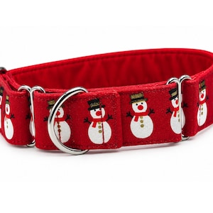 May include: A red fabric pet collar featuring a repeating pattern of white snowmen with black top hats, orange noses, and red scarves. The collar includes silver-toned metal hardware, including a D-ring for leash attachment and adjustable sliders.