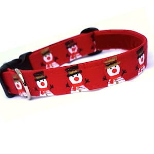 May include: A red fabric pet collar featuring a repeating pattern of snowmen wearing brown top hats and red scarves. The collar includes a black plastic side-release buckle and a silver-toned metal D-ring for attaching a leash or identification tag.