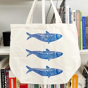 May include: Off-white canvas tote bag featuring three blue fish designs. The tote bag has two handles and is displayed against a backdrop of bookshelves filled with books. The fish are detailed with scales and fins.