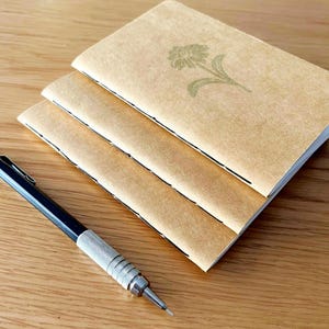 May include: Three kraft paper notebooks with a floral design and a black and silver mechanical pencil on a wooden surface. The notebooks are stacked, and the pencil is positioned diagonally across the scene.