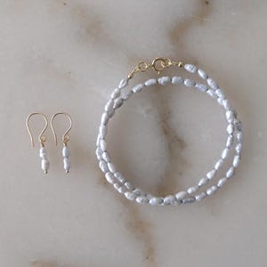 May include: A jewelry set consisting of a necklace and a pair of earrings. The necklace features a double strand of small, irregular, light gray pearls with a gold-toned clasp. The matching earrings have single pearl drops on gold-toned hooks.