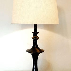 May include: A black table lamp with a cream-colored lampshade. The lamp has a glossy black base with a unique, tiered design. The lampshade is a neutral color, and the lamp is sitting on a wooden surface.
