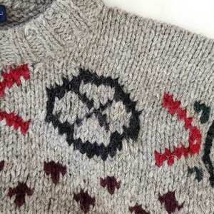 Peut inclure: Gros plan sur un pull en tricot gris avec un col montant. Le pull pr&eacute;sente un motif g&eacute;om&eacute;trique noir et rouge, ainsi que des accents bordeaux et verts. Le tricot est fait d'un fil &eacute;pais et textur&eacute;.