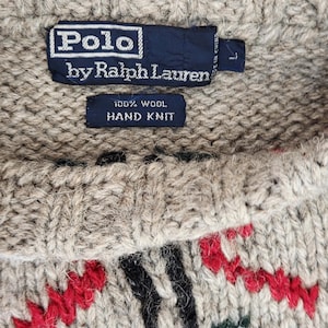 Peut inclure: Gros plan sur un pull en laine Polo by Ralph Lauren tricot&eacute; &agrave; la main. Le pull beige pr&eacute;sente un col ras du cou et un motif de motifs rouges, noirs et verts. L'&eacute;tiquette indique "100% Laine Tricot&eacute; &agrave; la Main."