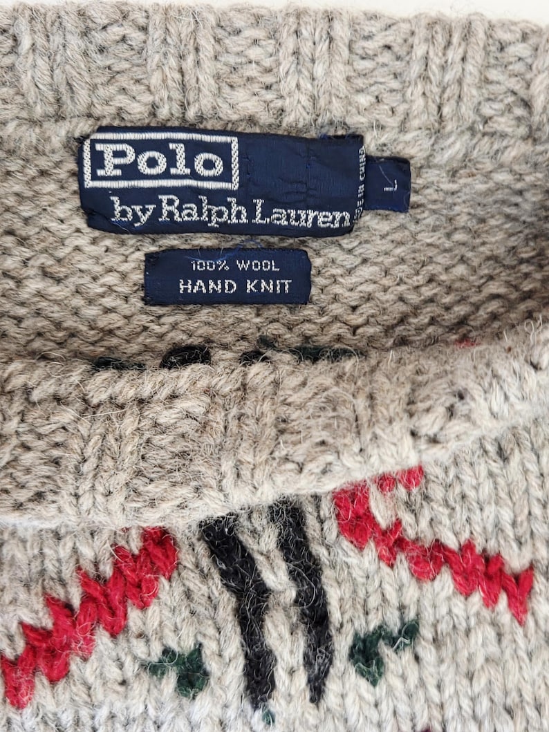 Peut inclure: Gros plan sur un pull en laine Polo by Ralph Lauren tricot&eacute; &agrave; la main. Le pull beige pr&eacute;sente un col ras du cou et un motif de motifs rouges, noirs et verts. L'&eacute;tiquette indique "100% Laine Tricot&eacute; &agrave; la Main."