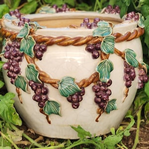 May include: White ceramic planter featuring a grapevine design. The planter is decorated with raised grapes and leaves in shades of green and purple.