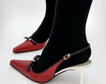 Puede incluir: Zapatos de tacón de cuero rojo con punta, correa de tobillo negra y tacón blanco pequeño. Los zapatos presentan un lazo negro con detalles dorados y lunares blancos. Se usan con medias negras.