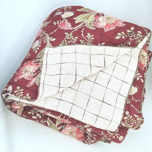 Könnte beinhalten: Eine rote und weiße Blumen-Steppdecke mit einem cremefarbenen karierten Muster auf der Rückseite. Die Steppdecke ist in der Hälfte gefaltet und die Blumen-Seite zeigt nach oben.