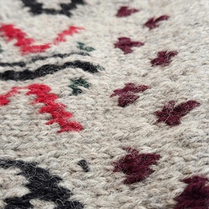Peut inclure: Gros plan sur un tissu tricot&eacute; beige avec un motif d&eacute;taill&eacute;. Le motif incorpore des formes florales et g&eacute;om&eacute;triques rouges, noires et bordeaux. La texture du tricot est visible, cr&eacute;ant un aspect douillet et textur&eacute;.