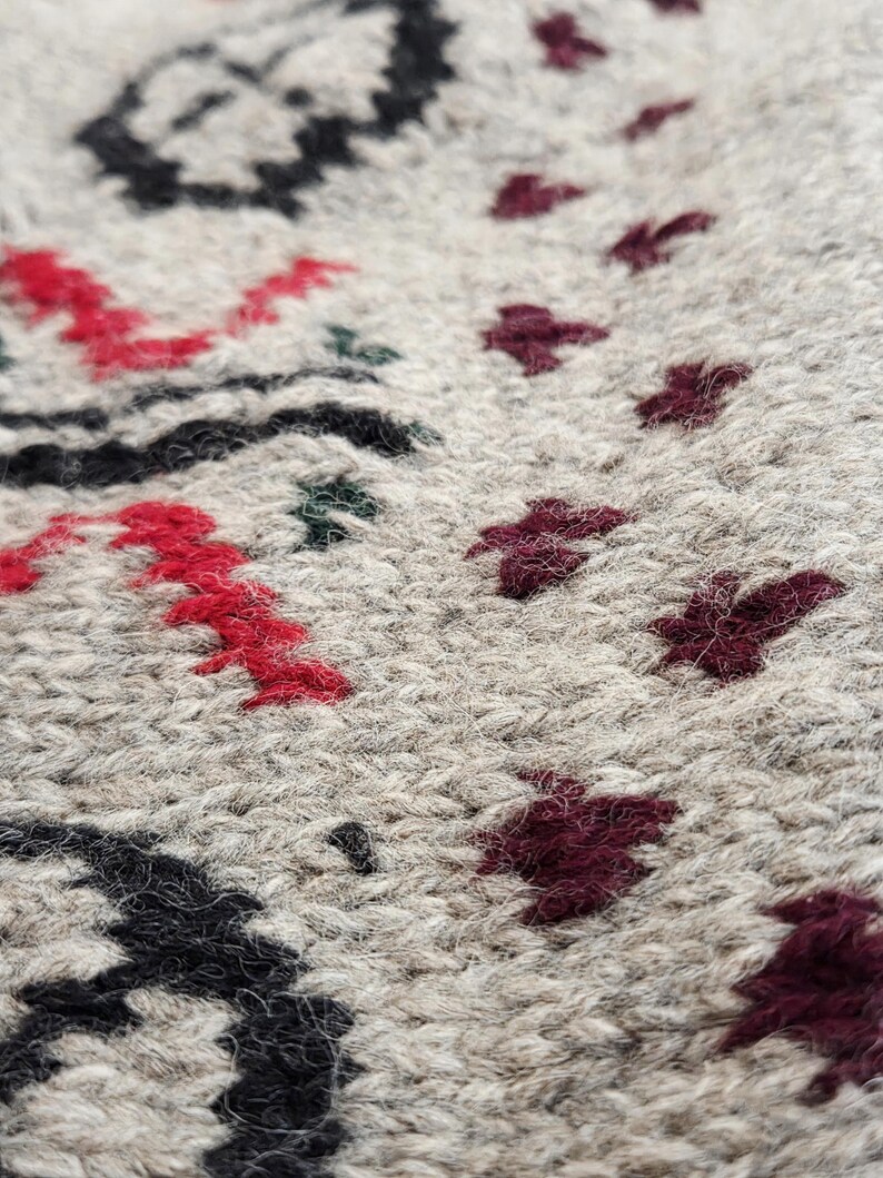 Peut inclure: Gros plan sur un tissu tricot&eacute; beige avec un motif d&eacute;taill&eacute;. Le motif incorpore des formes florales et g&eacute;om&eacute;triques rouges, noires et bordeaux. La texture du tricot est visible, cr&eacute;ant un aspect douillet et textur&eacute;.