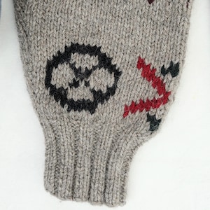 Peut inclure: Gros plan sur une manche tricot&eacute;e grise avec des motifs brod&eacute;s noirs et rouges. La manche pr&eacute;sente un embl&egrave;me circulaire noir et un motif angulaire rouge. Le tricot a un poignet c&ocirc;tel&eacute;.