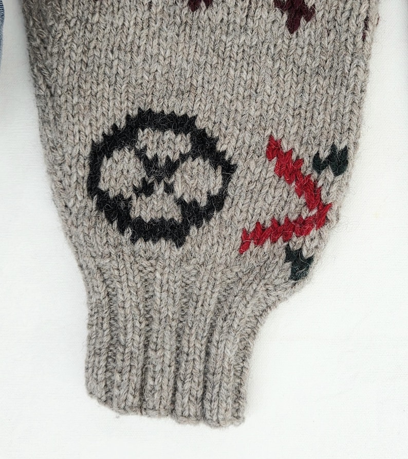 Peut inclure: Gros plan sur une manche tricot&eacute;e grise avec des motifs brod&eacute;s noirs et rouges. La manche pr&eacute;sente un embl&egrave;me circulaire noir et un motif angulaire rouge. Le tricot a un poignet c&ocirc;tel&eacute;.