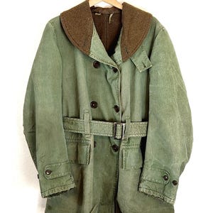Puede incluir: Una chaqueta verde de estilo militar con cuello de pana marrón, cierre de doble botonadura y cinturón. La chaqueta tiene dos bolsillos grandes en la parte delantera y dos bolsillos más pequeños en las mangas.