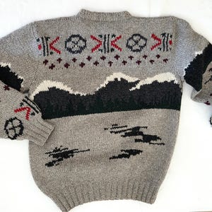 Peut inclure: Pull en tricot gris avec un motif de montagne et g&eacute;om&eacute;trique. Le pull pr&eacute;sente des accents noirs, blancs et rouges. Les manches ont des rayures noires et des motifs g&eacute;om&eacute;triques rouges et noirs. Un v&ecirc;tement confortable, chaud et &eacute;l&eacute;gant.