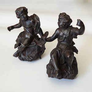 May include: Two antique bronze cherub sculptures. One cherub sits on a fish, the other on a rock. Both are seated, with one arm raised. The sculptures have a dark brown patina, typical of aged bronze.