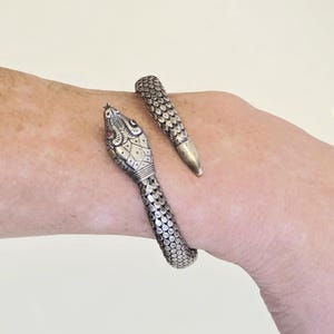 Puede incluir: Una pulsera de serpiente plateada con un patrón de escamas detallado. La cabeza de la serpiente presenta ojos de piedras preciosas rojas y marcas intrincadas. La pulsera está diseñada para envolverse alrededor de la muñeca, creando un accesorio único.