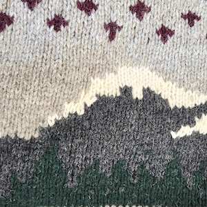 Peut inclure: Gros plan sur un pull en tricot gris avec un motif de montagne. Le pull pr&eacute;sente un paysage avec des &eacute;l&eacute;ments blancs, gris et verts. Des motifs g&eacute;om&eacute;triques rouges et bordeaux ornent le haut.