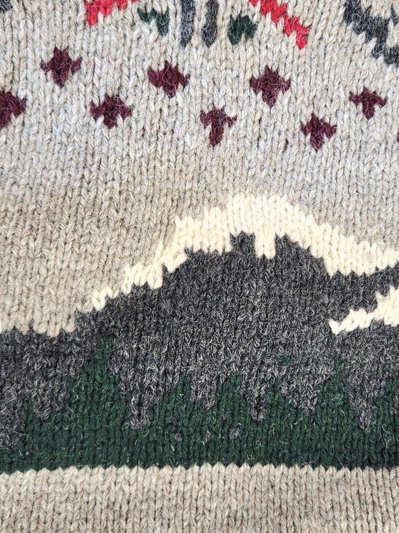 Peut inclure: Gros plan sur un pull en tricot gris avec un motif de montagne. Le pull pr&eacute;sente un paysage avec des &eacute;l&eacute;ments blancs, gris et verts. Des motifs g&eacute;om&eacute;triques rouges et bordeaux ornent le haut.