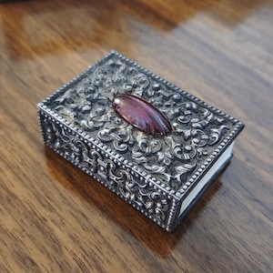 May include: A rectangular, antique silver box with ornate floral detailing. The box features a large, oval, reddish-purple gemstone on the lid. The box is likely used for storing small items, such as jewelry.
