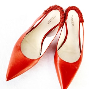 Puede incluir: Un par de zapatos de tacón de cuero naranja brillante con punta puntiaguda y diseño de slingback. Los zapatos tienen un tacón pequeño y el nombre de la marca "BOTTEGA VENETA" está impreso en el interior de cada zapato.