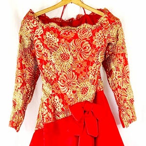 Puede incluir: Un vestido de terciopelo rojo con una superposición de encaje dorado y un gran lazo en la cintura.