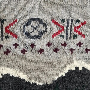 Peut inclure: Gros plan sur un pull en tricot gris avec des motifs g&eacute;om&eacute;triques rouges, vert fonc&eacute; et noirs. Le pull pr&eacute;sente une encolure ras du cou et un motif textur&eacute;, avec un motif de montagne en bas.