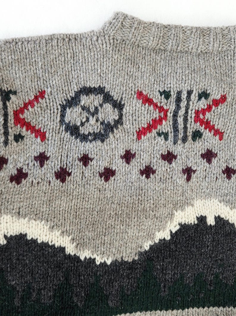 Peut inclure: Gros plan sur un pull en tricot gris avec des motifs g&eacute;om&eacute;triques rouges, vert fonc&eacute; et noirs. Le pull pr&eacute;sente une encolure ras du cou et un motif textur&eacute;, avec un motif de montagne en bas.