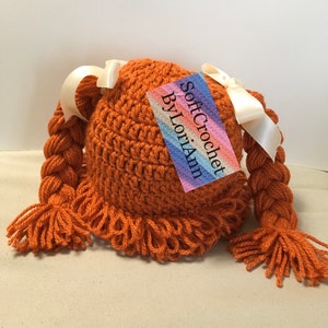 May include: A crocheted hat with two long braids, each with a white ribbon tied at the top. The hat is a rust orange color and has a fringe around the bottom. The hat is labeled "Soft Crochet by LoriAnn".